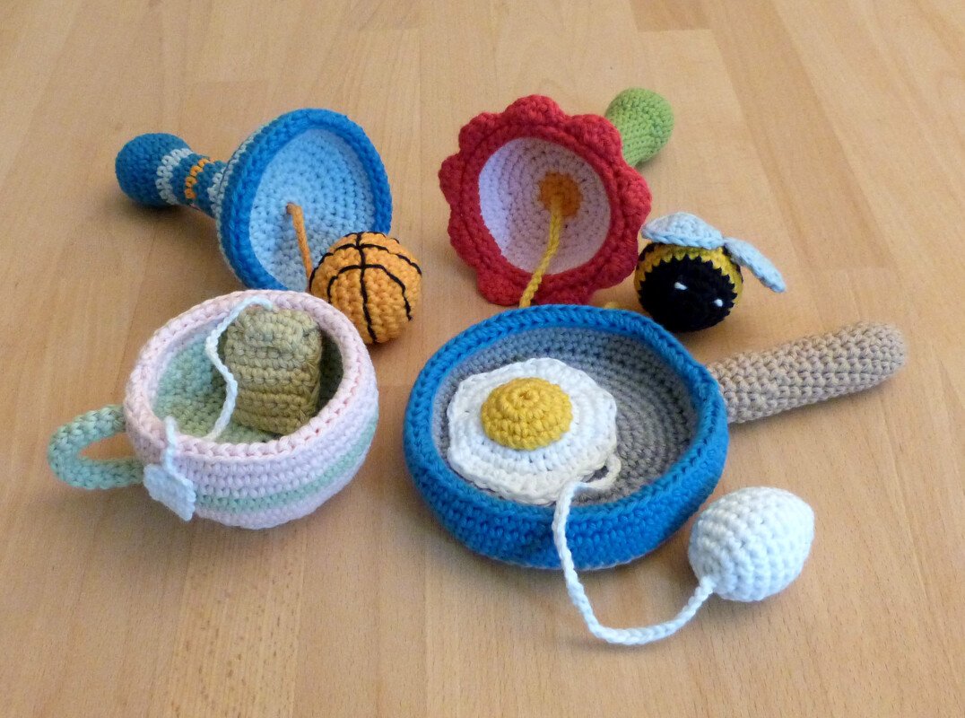 Gehäkelte Kinderspielzeuge: blaue Pfanne mit Spiegelei, pastellfarbene Tasse, blaue Trichterrassel, kleiner Basketball, Blumenrassel und Bienenrassel auf Holztisch