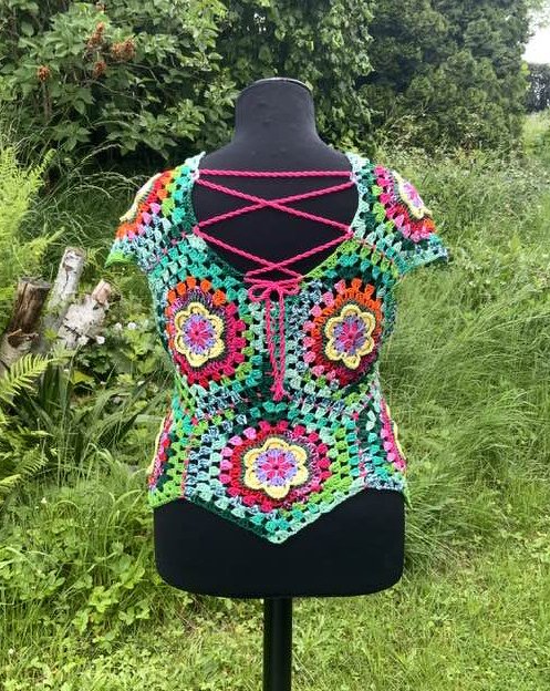 Buntes Häkeltop aus Granny Squares auf schwarzer Schneiderpuppe vor grünen Pflanzen, mit pinker Kreuzschnürung am Rücken.
