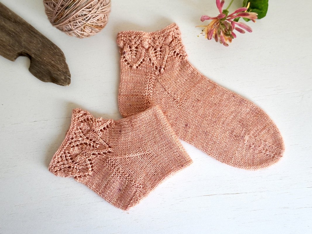 Paar roséfarbener gestrickter Socken mit Spitzenbündchen und Wollknäuel auf hellem Untergrund