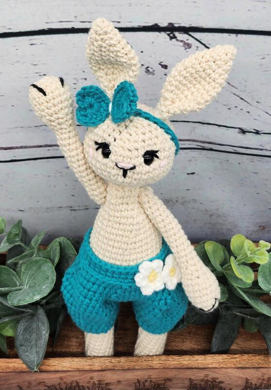 Gehäkeltes Amigurumi-Hasenpüppchen in Creme mit türkisfarbener Hose und Schleife, ein Arm erhoben