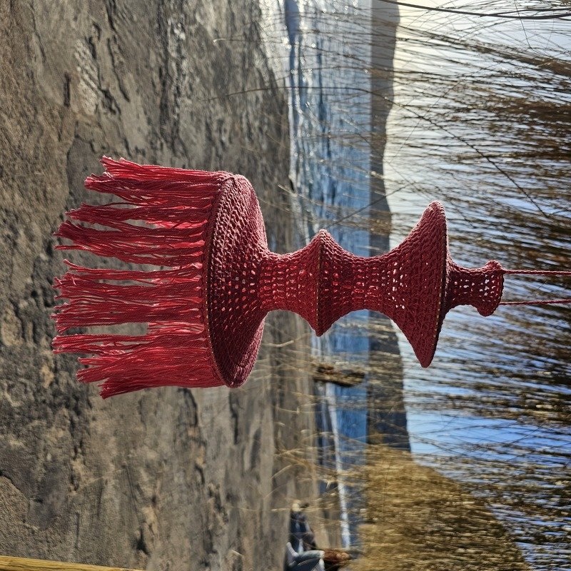 Red crocheted hanging pendant with tiered shape and long fringe suspended outdoors by the water