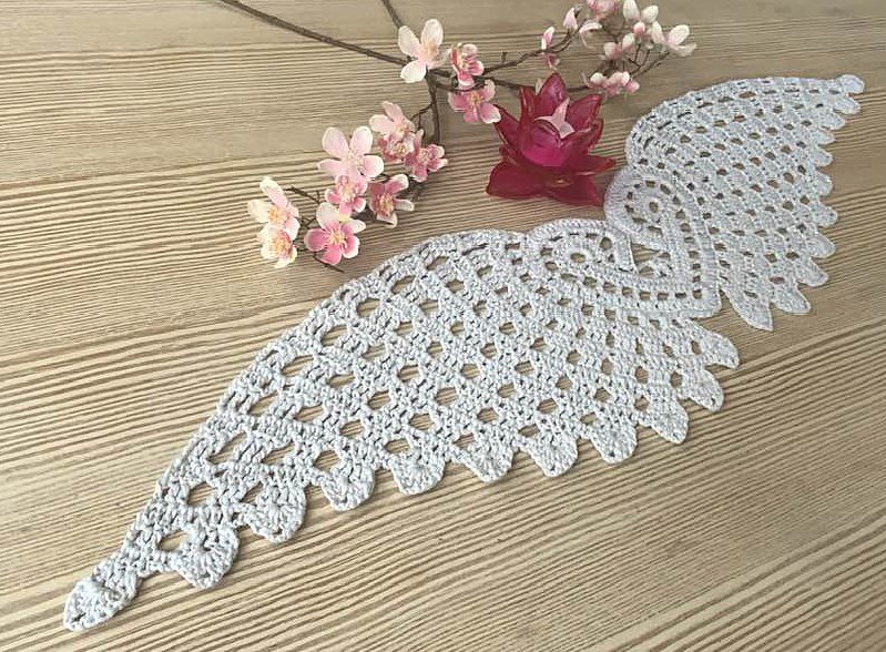 Hellgraues gehäkeltes Deckchen in Engelsflügelform auf Holz mit rosa Blüten und Teelicht