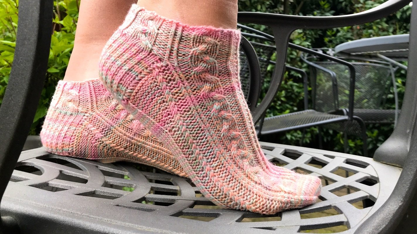 Füße in gestrickten kurzen Socken mit Zopfmuster in Rosa- und Grüntönen auf einem Metallstuhl