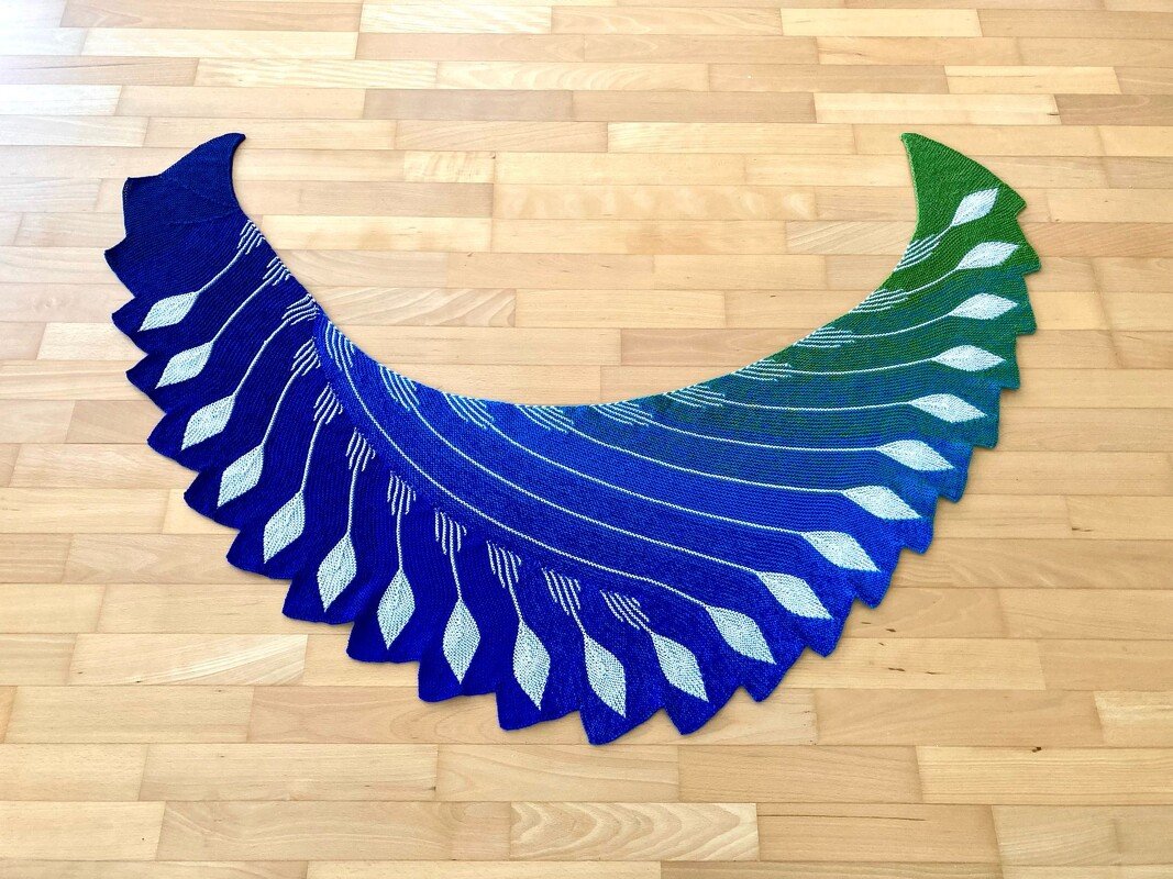 Gestrickter halbmondförmiger Schal mit blau-grünem Farbverlauf und weißen Federmotiven auf hellem Holzboden