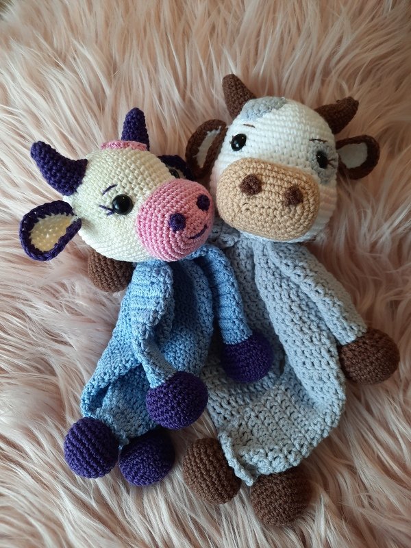 Zwei handgehäkelte Kuh‑Schmusetücher mit Amigurumi‑Köpfen auf rosafarbenem Plüschuntergrund