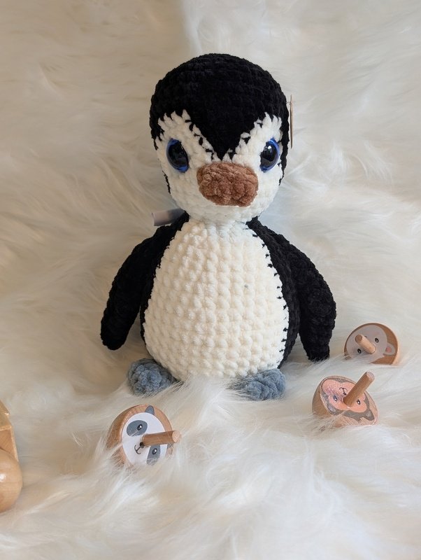 Gehäkeltes Pinguin‑Amigurumi mit blauen Augen und braunem Schnabel auf weißem flauschigem Untergrund, herum liegen hölzerne Kreiseln.