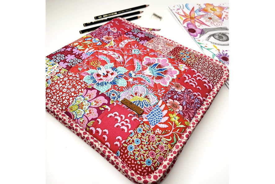 Gesteppte Patchwork-Tasche in Rottönen mit floralen Mustern, liegend neben Bleistiften und einer Zeichnung.