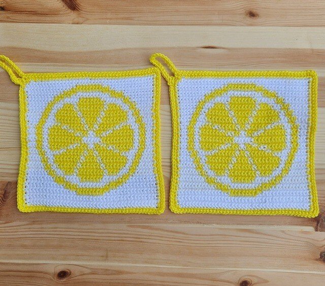 Zwei quadratische gehäkelte Topflappen in Weiß und Gelb mit Zitronenscheibenmotiv auf Holzoberfläche.