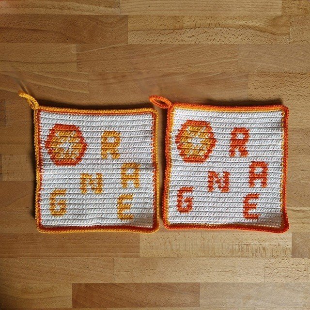 Zwei weiße gehäkelte Topflappen mit orangefarbenem 'ORANGE'-Schriftzug und stilisierter Orangenscheibe auf Holz.