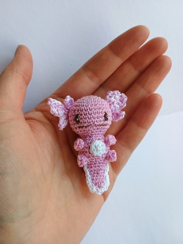 Tiny pink crocheted axolotl amigurumi with a white belly and tail edge, resting on an open hand.