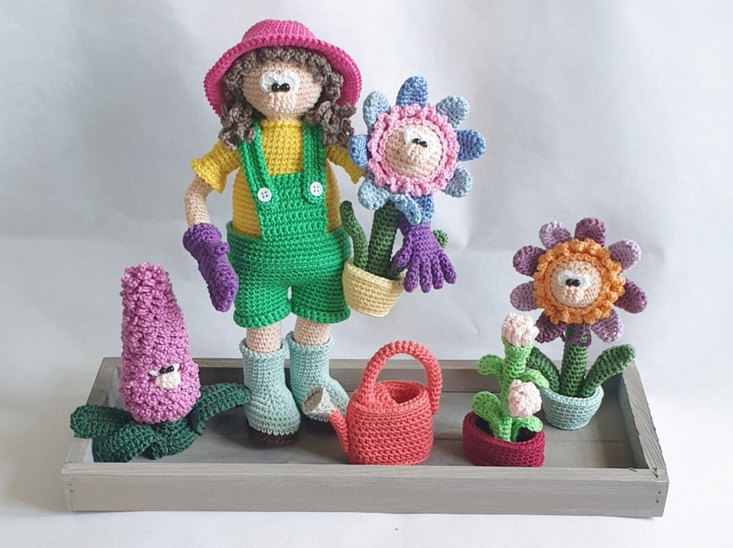 Gehäkelte Amigurumi-Gärtnerin im rosa Hut und grüner Latzhose, mit Topfblumen und roter Gießkanne auf einem Holztablett.