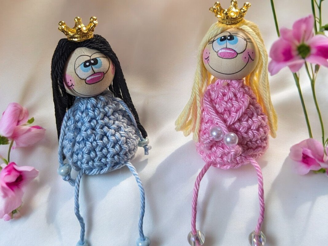 Zwei gehäkelte Prinzessinnenpuppen in Blau und Rosa mit Kronen und Perlen auf hellem Untergrund