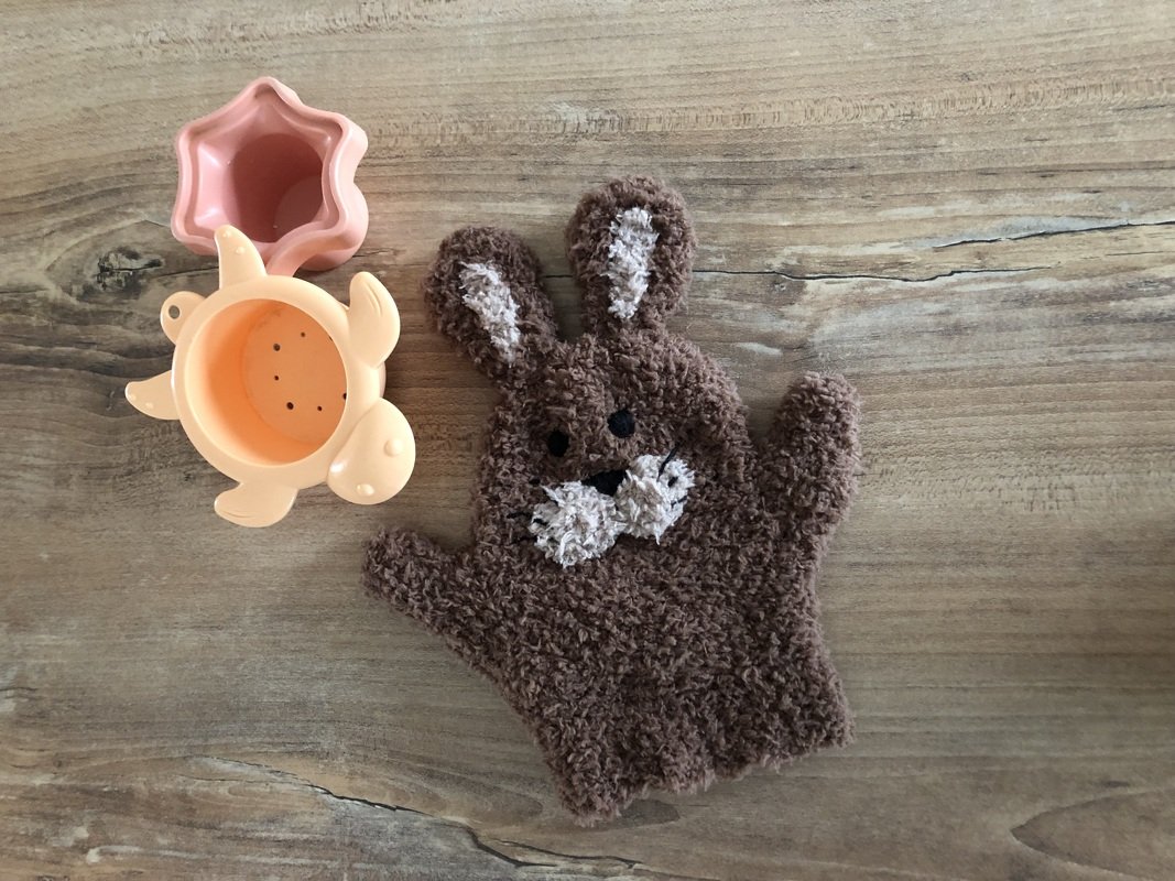 Brauner, flauschiger Hasen-Waschlappen mit weißen Gesichts- und Ohrdetails neben rosa Sternförmchen und orangefarbener Schildkrötenform auf Holz.