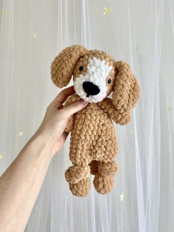 Hand holding a light brown and white crochet amigurumi dog with floppy ears against a sheer star-patterned curtain.
