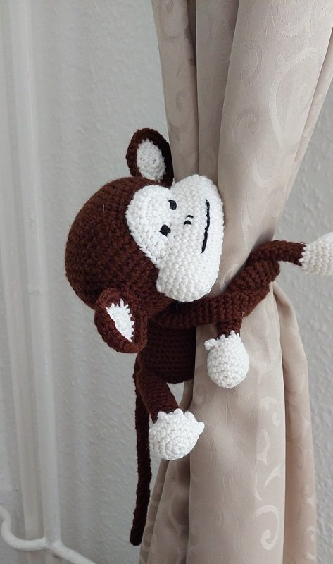 Gehäkelter braun-weißer Amigurumi-Affe umschlingt eine helle Gardine als Raffhalter.