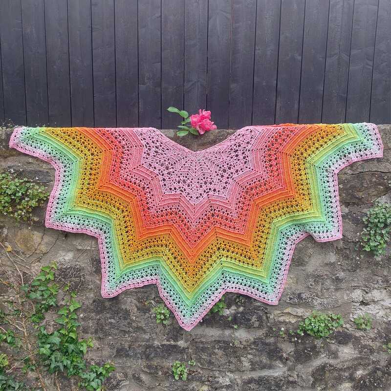 Star-shaped crochet shawl with pink-to-green gradient, draped on a stone wall with a pink rose