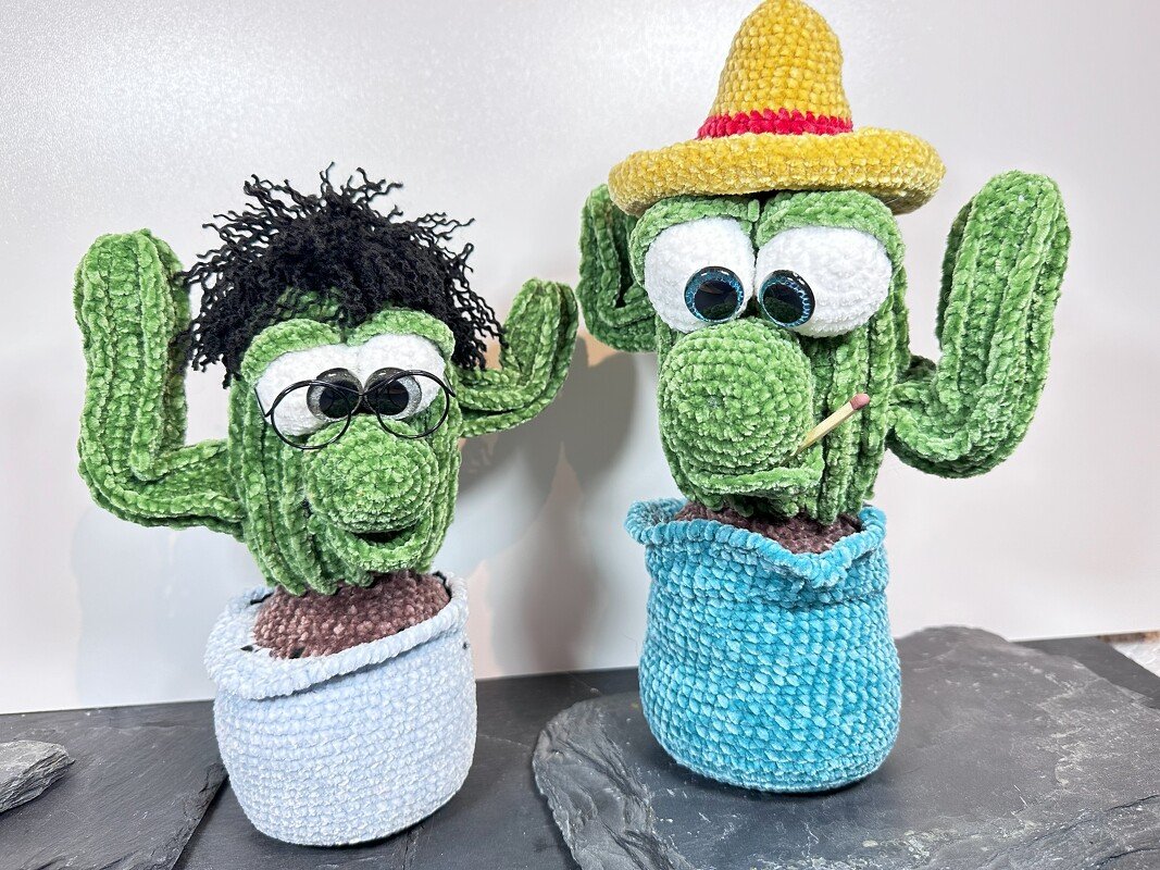 Zwei grüne gehäkelte Amigurumi-Kakteen in Stofftöpfen, einer mit Drahtbrille und schwarzem Garnhaar, der andere mit gelbem Hut und Streichholz