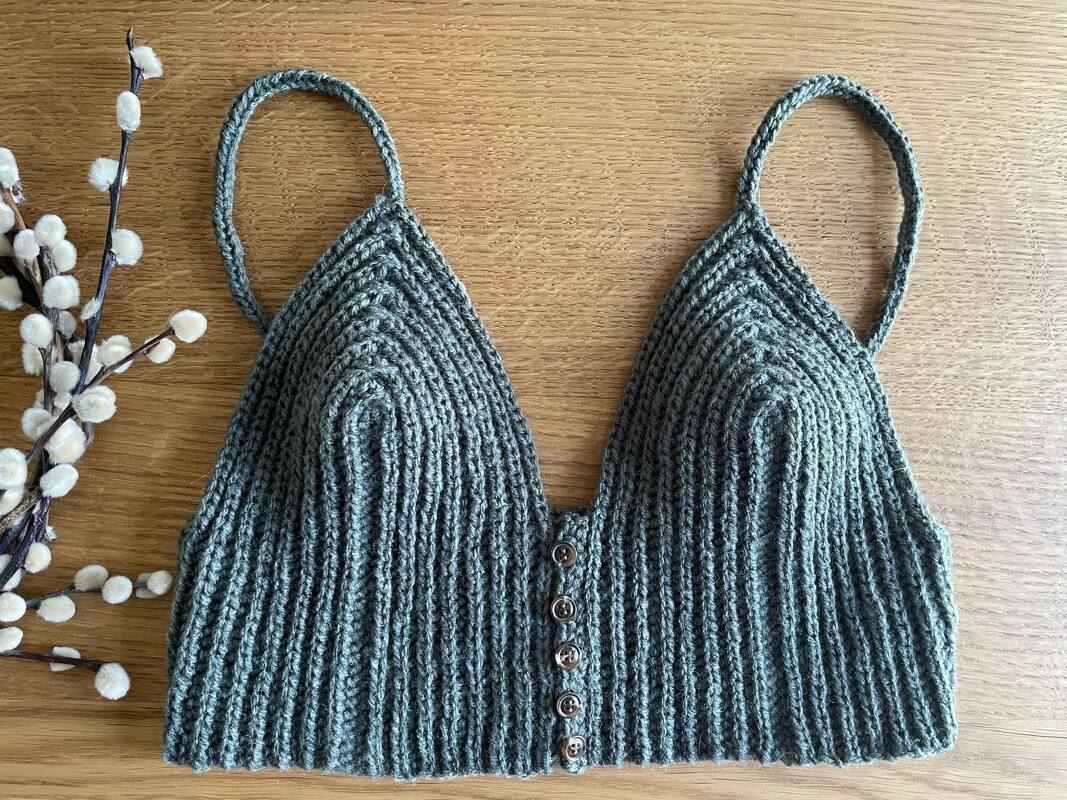 Gehäkeltes dunkelgrünes Crop-Top mit Rippenmuster, schmalen Trägern und mittiger Knopfleiste auf Holz, dekoriert mit Weidenkätzchen.