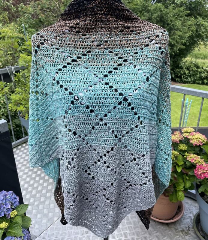 Drapiertes gehäkeltes Dreieckstuch mit Farbverlauf von Dunkelbraun/Grau über Türkis zu Hellgrau und Rautenmuster, auf Balkon mit Pflanzen im Hintergrund