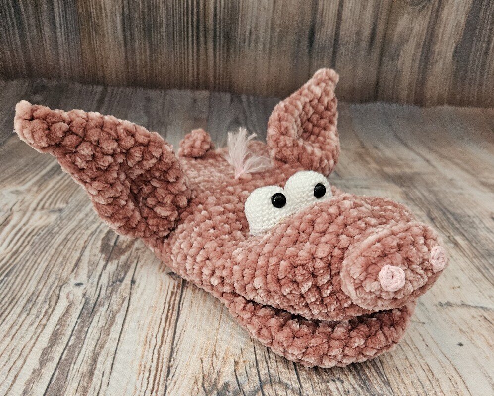 Rosa gehäkeltes Amigurumi in Schweineform mit großen Ohren und aufgesetzter Schnauze, liegend auf Holzboden