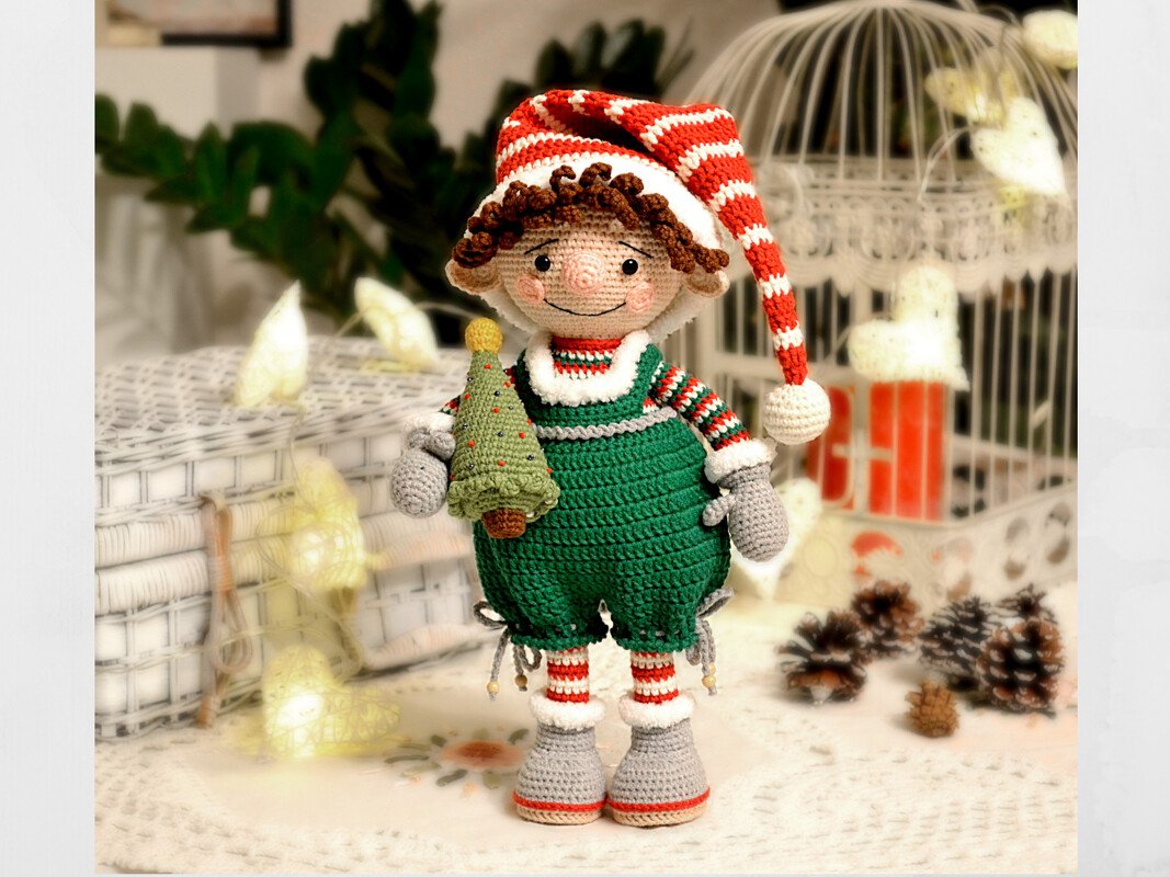 Crocheted amigurumi elf in green overalls and a red-and-white striped hat holding a small Christmas tree.
