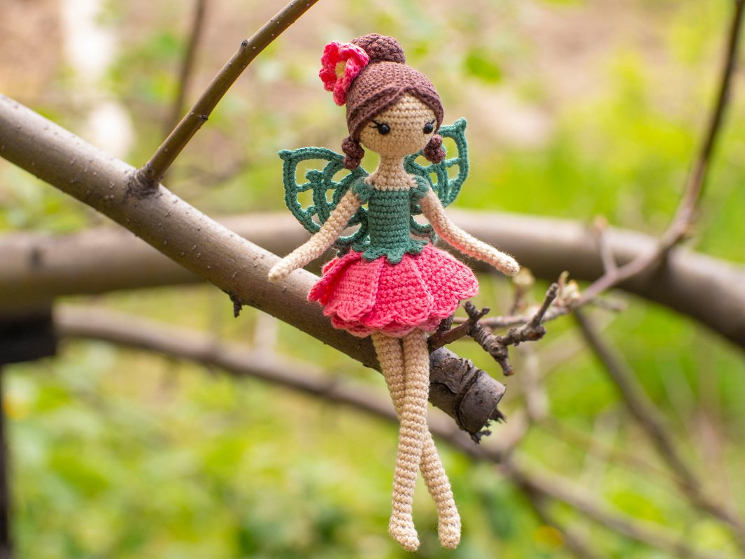 Handmade crocheted fairy amigurumi doll with green wings and pink skirt sitting on a branch