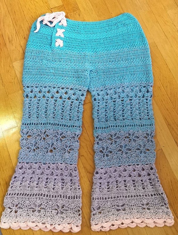 Gehäkelte Damenhose in Türkis bis Lila mit offenem Muster und Muschelsaum, liegend auf Holzboden