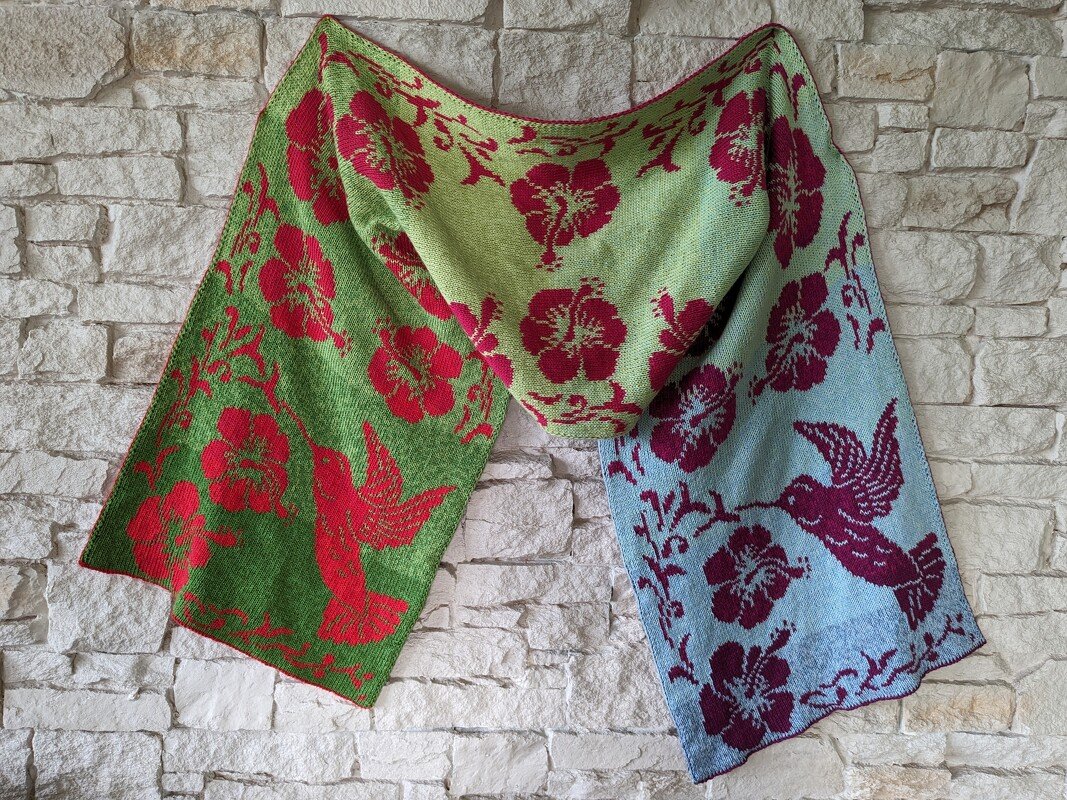 Rectangular knitted shawl draped on a stone wall, left panel green with red bird and flower motifs, right panel pale blue with maroon motifs.