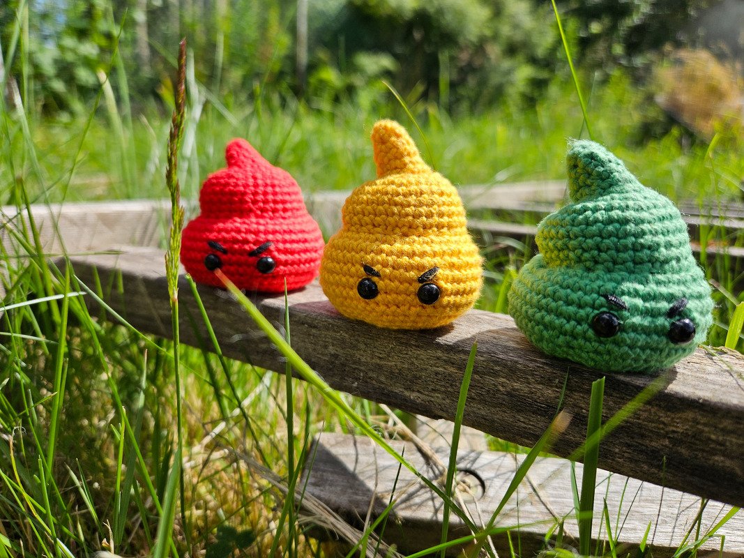 Drei gehäkelte Amigurumi-Emojis (rot, gelb, grün) mit mürrischem Gesicht auf einem Holzbrett im Gras.