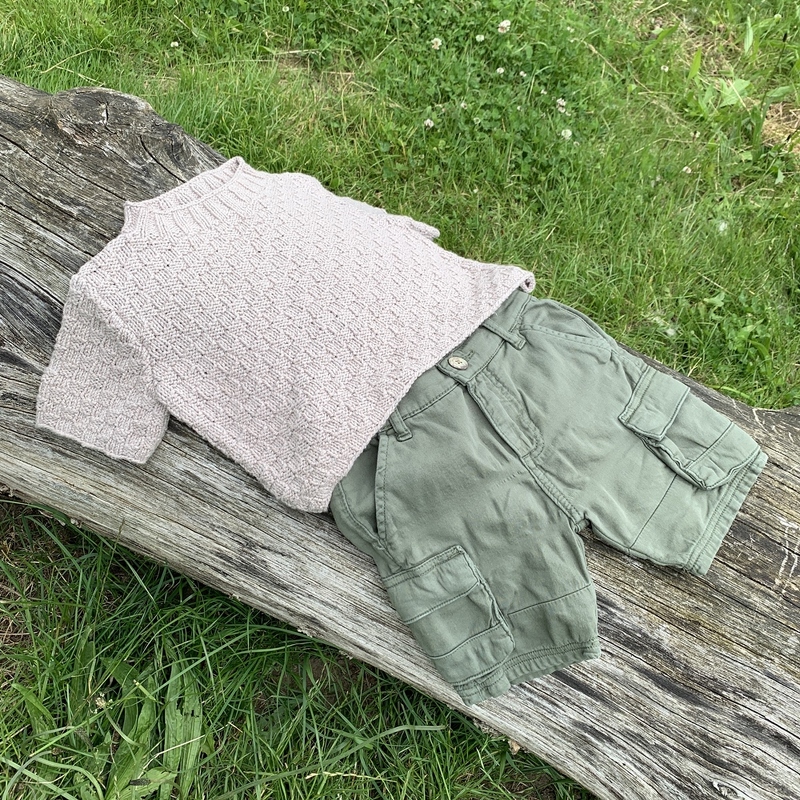 Zartrosa gestrickter kurzärmeliger Kinderpullover mit Stehkragen neben olivgrünen Cargo-Shorts auf einem Holzstamm