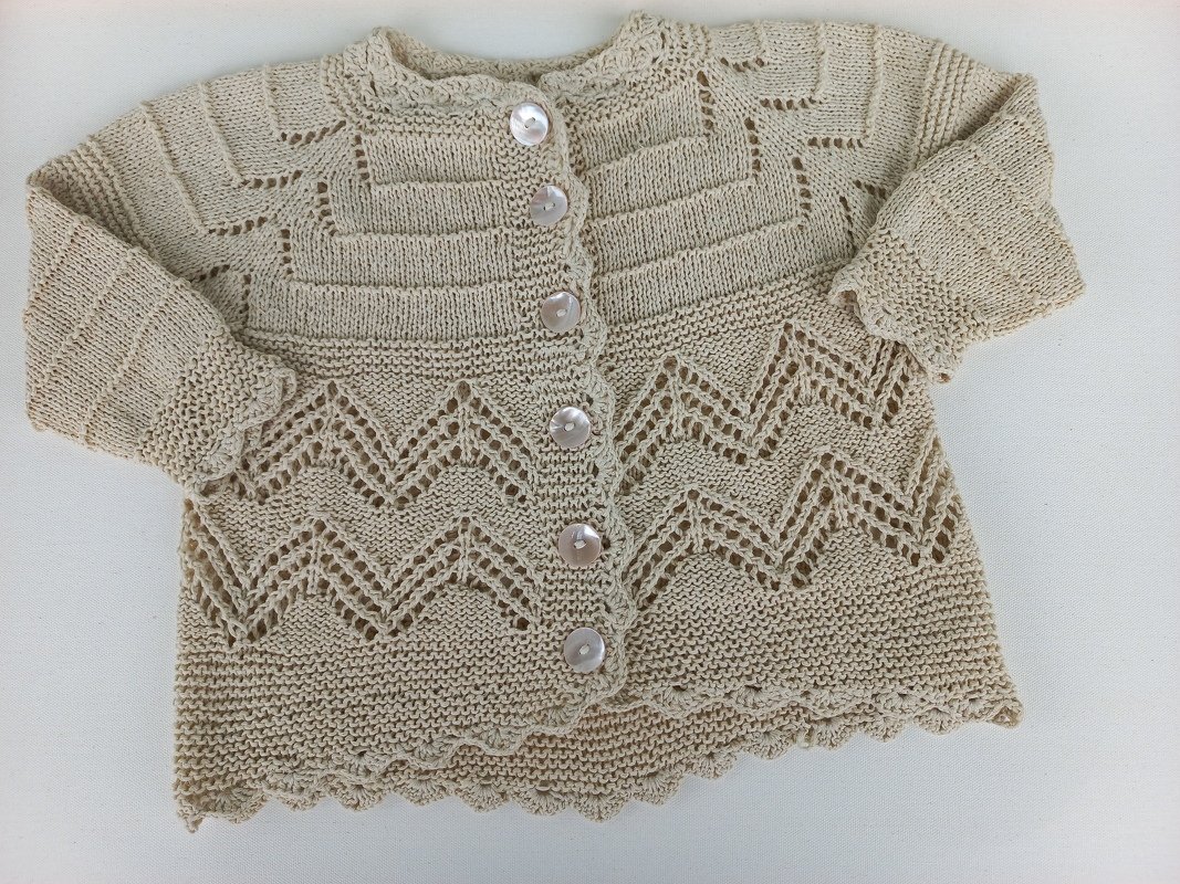Beige gestrickte Babyjacke mit Zickzack-Lochmuster, Rippenstruktur oben und fünf Perlmuttknöpfen, flach auf hellem Hintergrund.