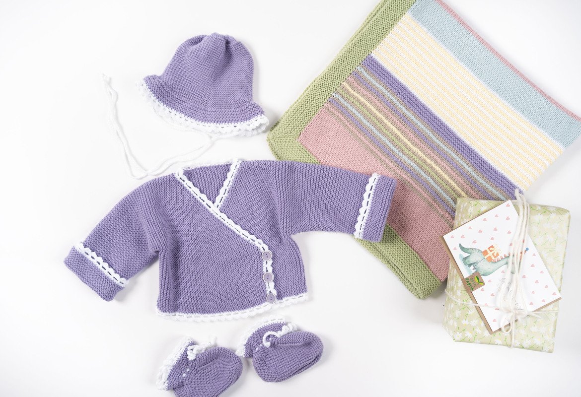 Lavendelfarbene Wickeljacke, Mütze und Schühchen mit weißer Kante neben einer pastellgestreiften Babydecke und einem verpackten Geschenk mit Karte.