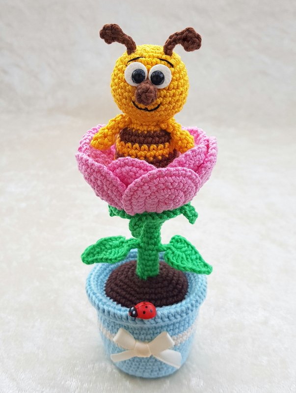 Nahaufnahme: gehäkelte Biene mit braunen Streifen in rosa Blüte auf grünem Stiel in blauem Topf mit Schleife und Marienkäfer-Button