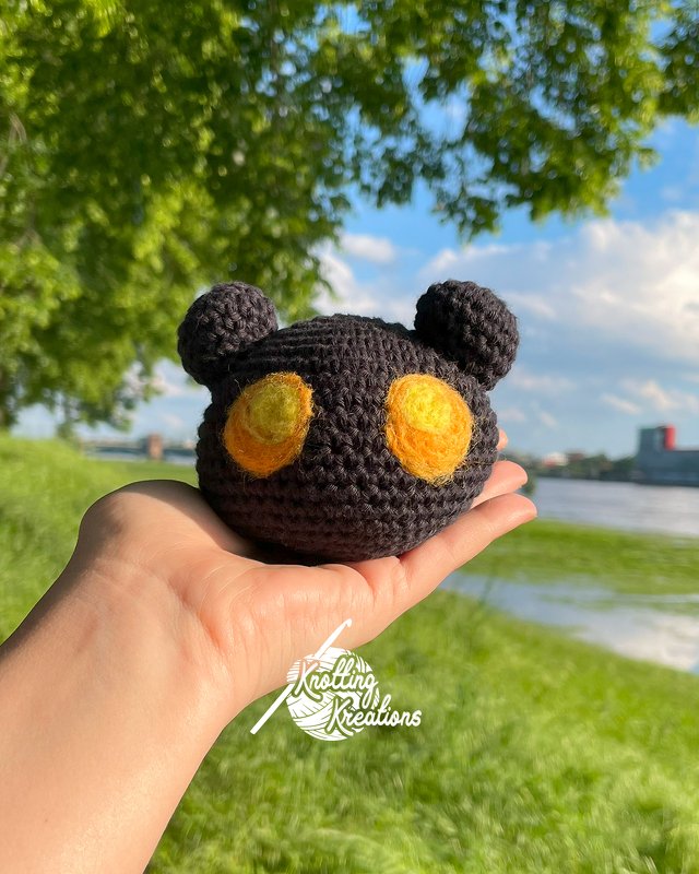 Hand präsentiert ein dunkles, rundes Amigurumi‑Köpfchen mit großen gelben Augen vor Fluss und Grün.