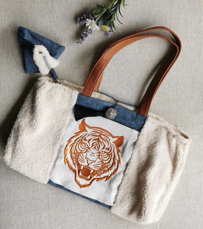Beigefarbene Sherpa-Tasche mit Jeansstoff und orangefarbener Tigerkopf-Stickerei auf der Vorderseite; braune Träger und ein kleiner Reißverschlussbeutel daneben.