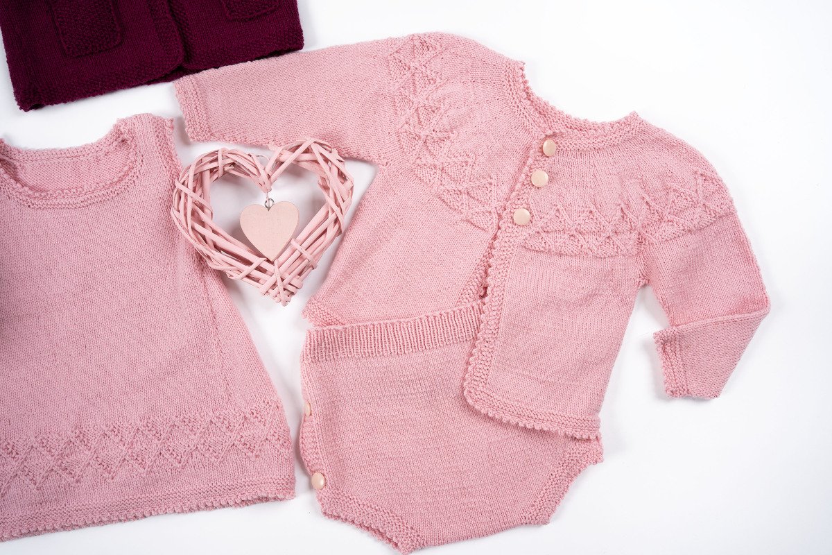 Rosa gestricktes Baby-Set mit Jacke, Strampler und ärmellosem Oberteil, feines Ajourmuster und geflochtenes Herz als Dekoration auf hellem Hintergrund