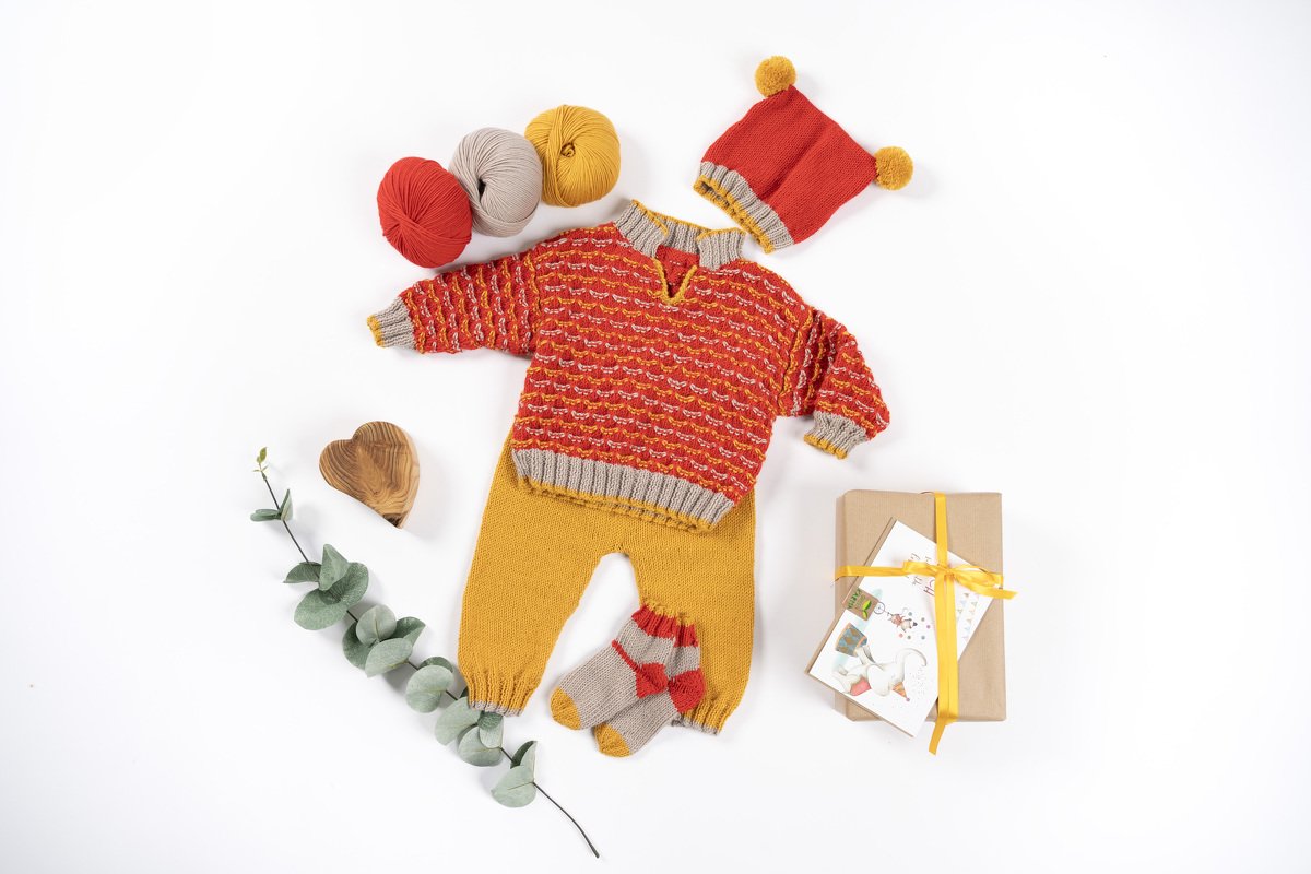 Gestricktes Babyset: roter Pullover, senfgelbe Hose, Mütze mit Bommel, kleine Socken, Garnknäuel und Geschenkbox
