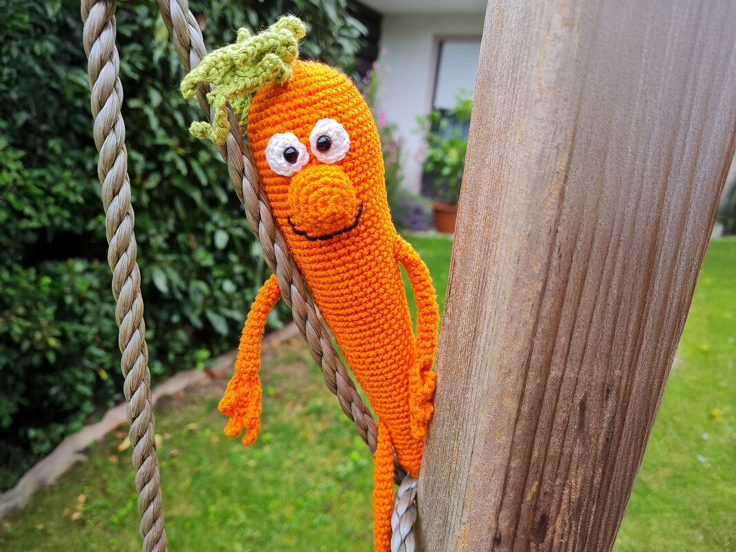 Hand-crocheted orange carrot amigurumi with embroidered eyes and green leaf top hanging on a rope