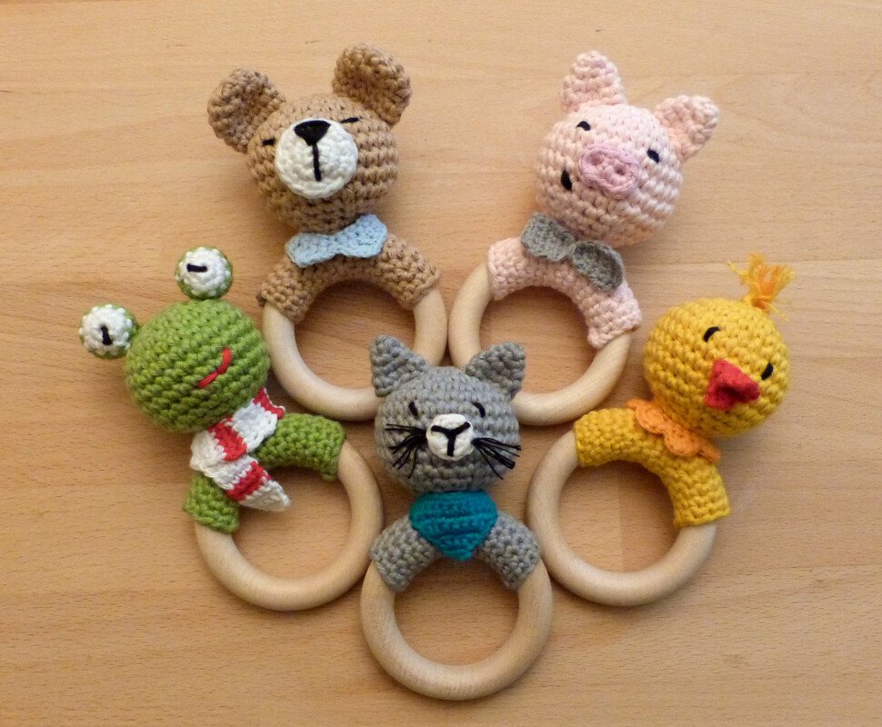 Fünf gehäkelte Baby-Greiflinge mit Holzringen: Bär, Schwein, Frosch, Katze und Küken.