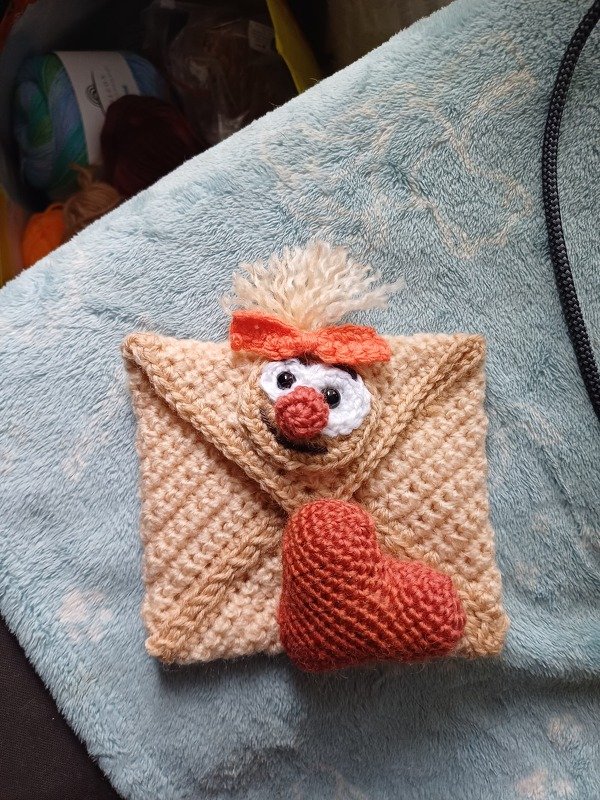 Beigefarbener gehäkelter Umschlag mit Amigurumi-Gesicht, oranger Schleife und rotem Herz auf hellblauem Stoff.