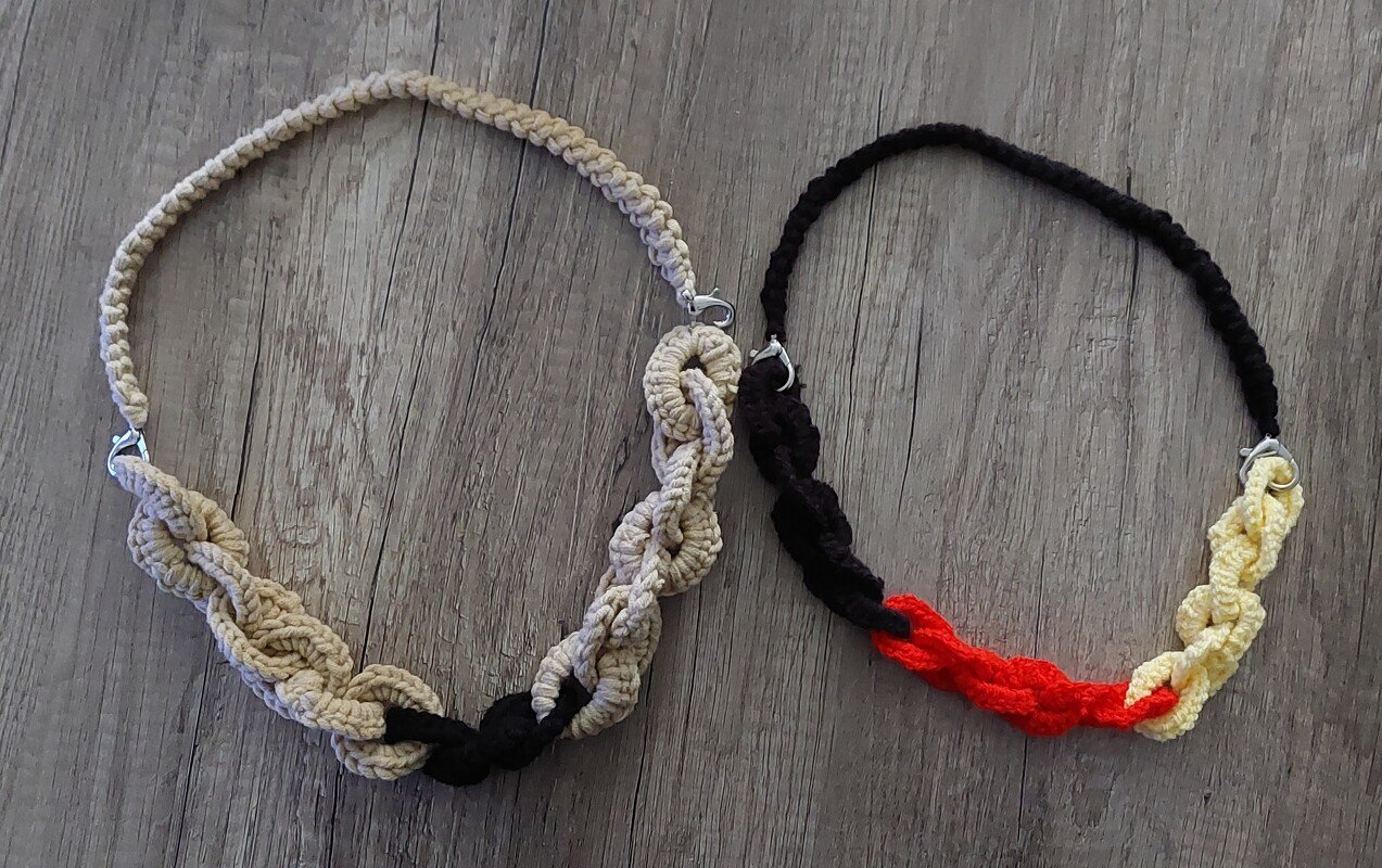 Zwei gehäkelte Ketten im Kettendesign, eine beige‑schwarz, eine schwarz‑rot‑gelb, auf Holz.
