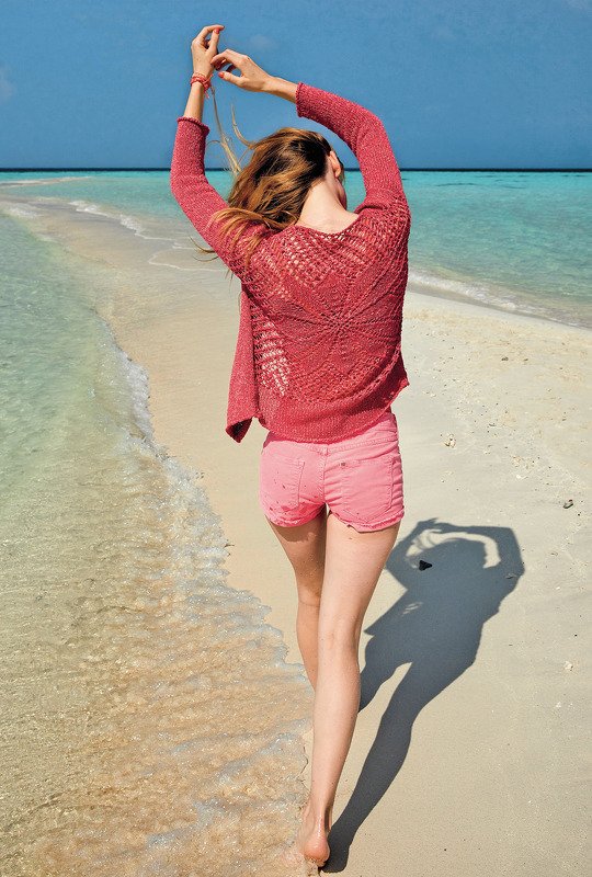 Rückenansicht: Frau in korallroter, leicht gestrickter Jacke mit Ajourmuster und Shorts am Strand