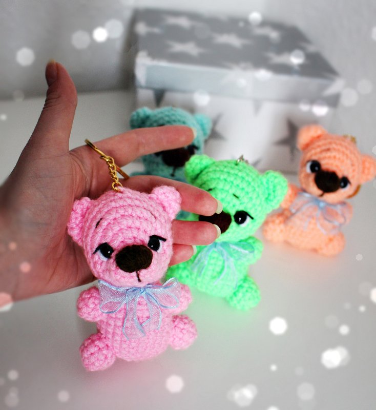 Mehrere gehäkelte Teddybär-Schlüsselanhänger in Rosa, Mintgrün, Türkis und Apricot, einer wird von einer Hand gehalten.