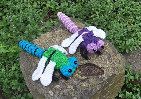 Zwei farbenfrohe Amigurumi‑Libellen (grün‑blau und lila‑rosa) auf einem Stein.