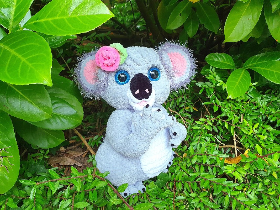 Gray crocheted koala amigurumi with big blue eyes and a pink flower on its head, sitting surrounded by green leaves