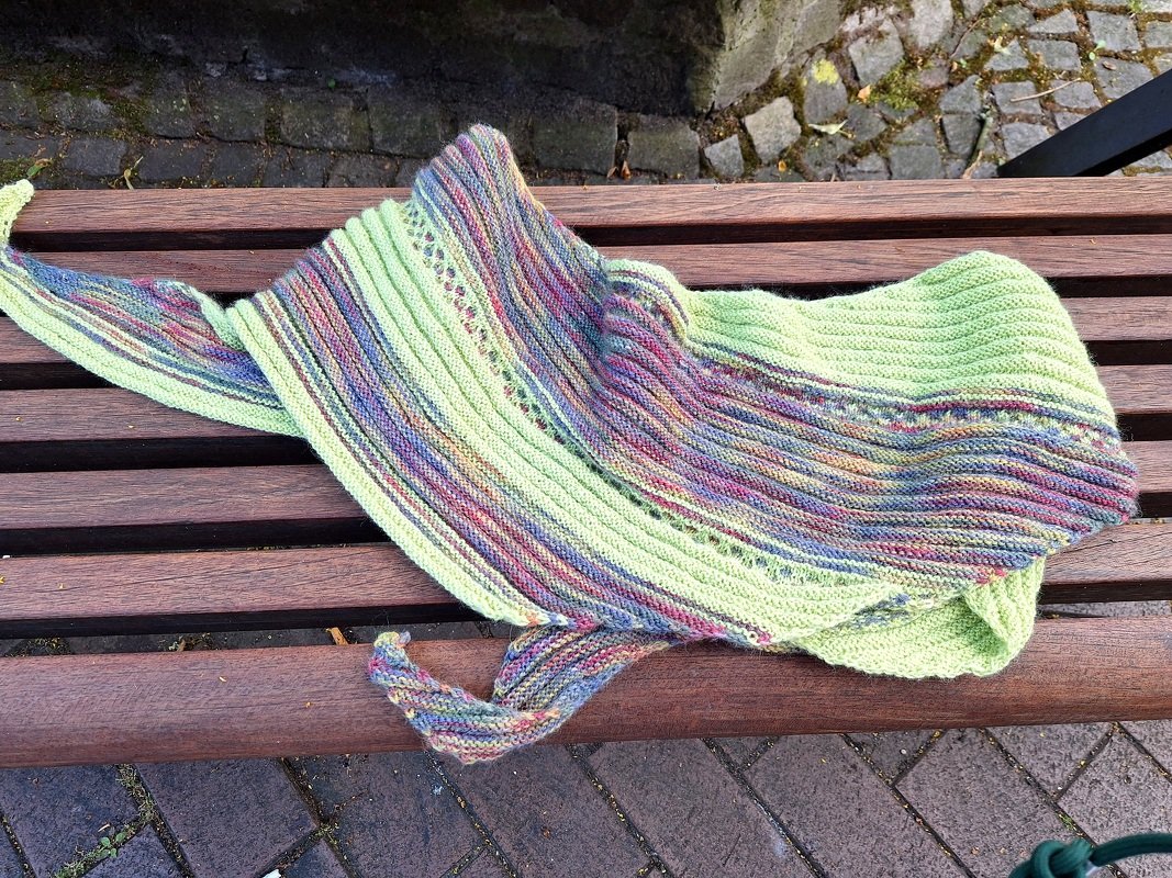 Asymmetrisches gestricktes Dreieckstuch in Hellgrün und mehrfarbig mit Rippen und Lochreihe auf Holzbank