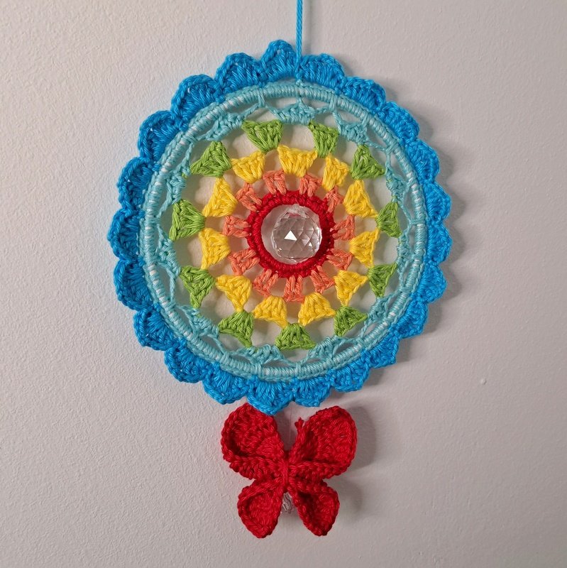 Gehäkelter runder Traumfänger in Regenbogenfarben mit facettiertem Kristall in der Mitte und rotem Schmetterling an weißer Wand