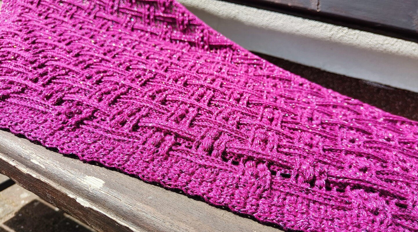 Close-up of fuchsia crochet triangle shawl with raised openwork stitch, ribbed border, and sparkly yarn on a wooden bench