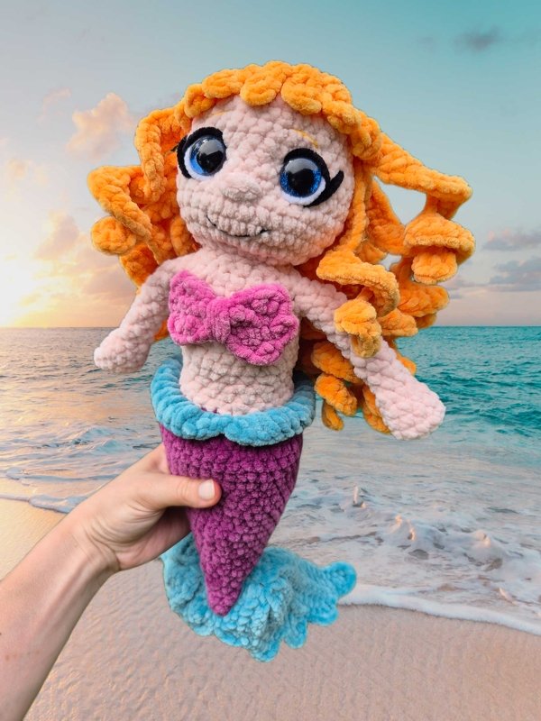 Hand hält gehäkelte Meerjungfrau-Amigurumi mit orangen Locken und violettem Schwanz am Strand