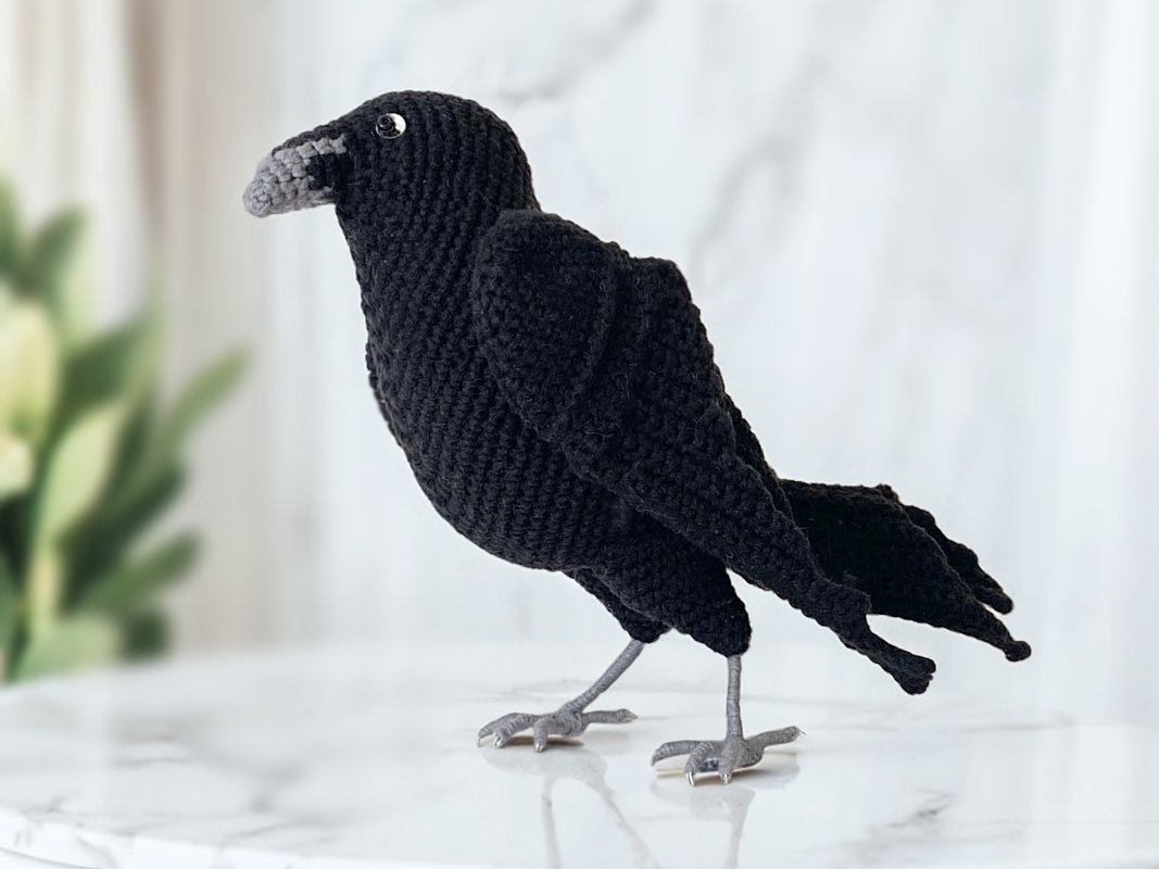 Black crocheted crow amigurumi with grey beak and grey legs standing on a marble surface